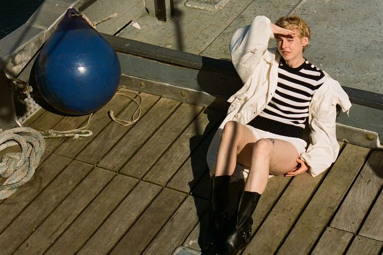 Singer-songwriter Fenne Lily sitting on a wooden boat deck in a striped top and white jacket.